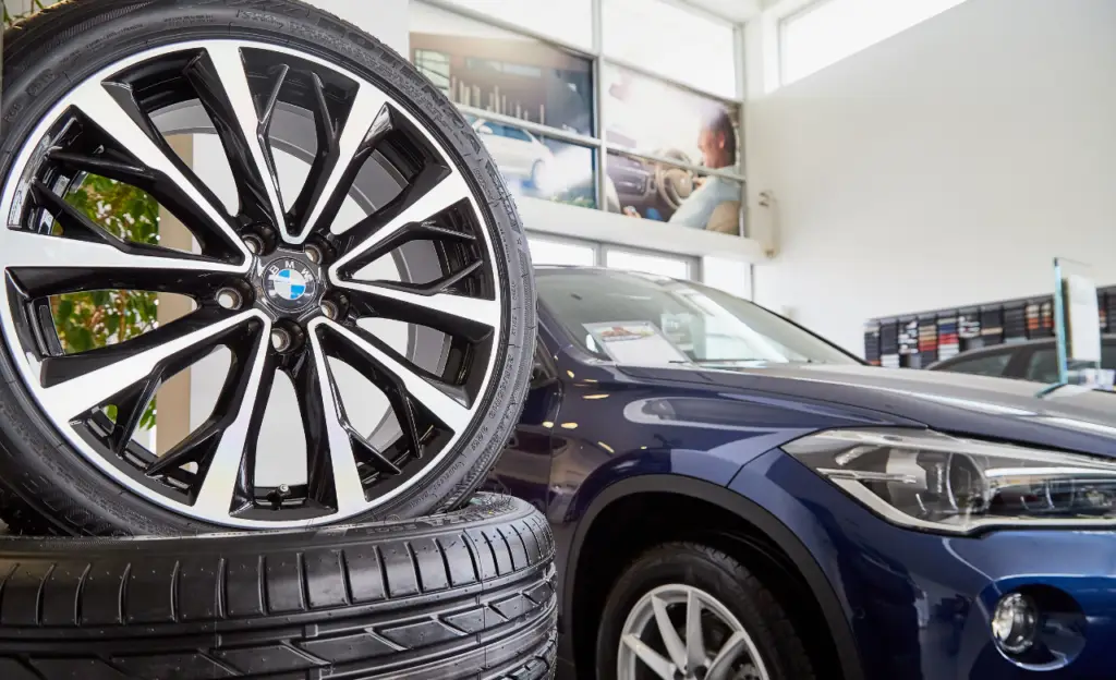 Close-up of a stylish car wheel with a BMW logo, displayed on a stack of tires. A sleek blue BMW vehicle is blurred in the background, inside a well-lit showroom with car-related posters on the walls. EuroMechanic | Top Reviewed Auto Repair Shop | Greater Toronto and Guelph | Visit Euromechanic