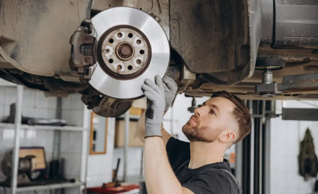 A mechanic wearing gloves is inspecting a brake disc on a lifted car in a workshop. He is focused, with tools and shelves visible in the background. EuroMechanic | Top Reviewed Auto Repair Shop | Greater Toronto and Guelph | Visit Euromechanic