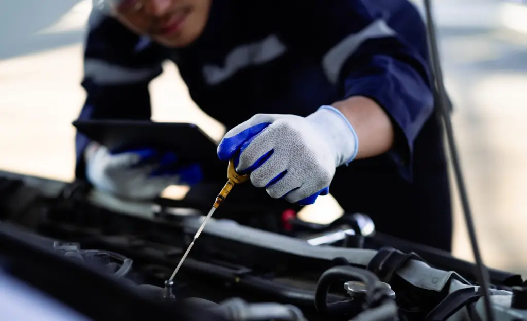 A mechanic wearing a blue and black uniform and white gloves checks a cars oil level with a dipstick while holding a tablet in the other hand. The cars hood is open, revealing parts of the engine. EuroMechanic | Top Reviewed Auto Repair Shop | Greater Toronto and Guelph | Visit Euromechanic