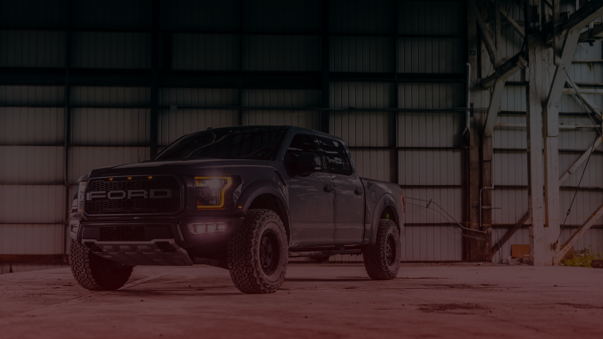 A black Ford Raptor truck, meticulously inspected by a skilled Ford mechanic in Greater Toronto, is parked inside a dimly lit industrial warehouse. Its lights gleam, revealing rugged off-road tires amid large metal beams and a hint of open space. EuroMechanic | Top Reviewed Auto Repair Shop | Greater Toronto and Guelph | Visit Euromechanic