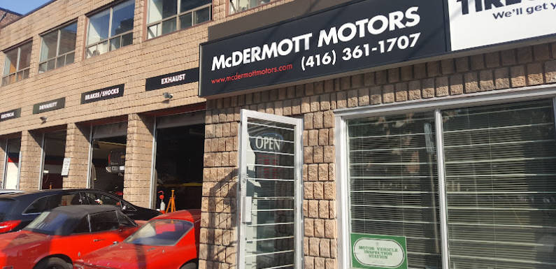Exterior view of McDermott Motors, a trusted Euro Mechanic location, with an open sign on the door. A red car is parked outside the garage, beneath signs for exhaust, brakes, shocks, tires, and rims above the service bays. Contact number is prominently displayed on the facade. EuroMechanic | Top Reviewed Auto Repair Shop | Greater Toronto and Guelph | Visit Euromechanic
