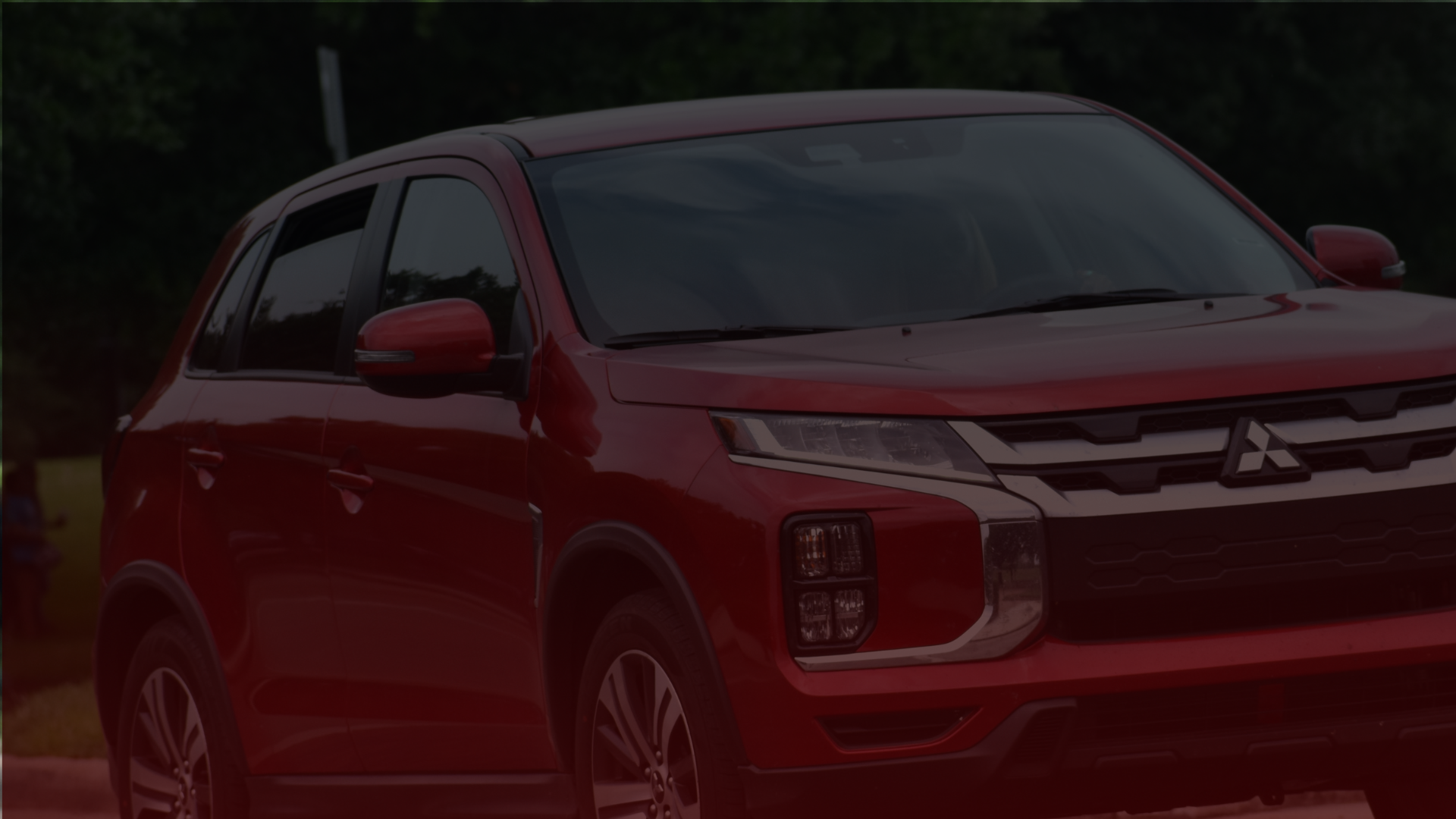 A red Mitsubishi SUV is parked outdoors, awaiting its next adventure around Greater Toronto. The cars design features a sleek grille, LED headlights, and side mirrors matching the body color. In the background is a blurred view of green trees under a cloudy sky. EuroMechanic | Top Reviewed Auto Repair Shop | Greater Toronto and Guelph | Visit Euromechanic