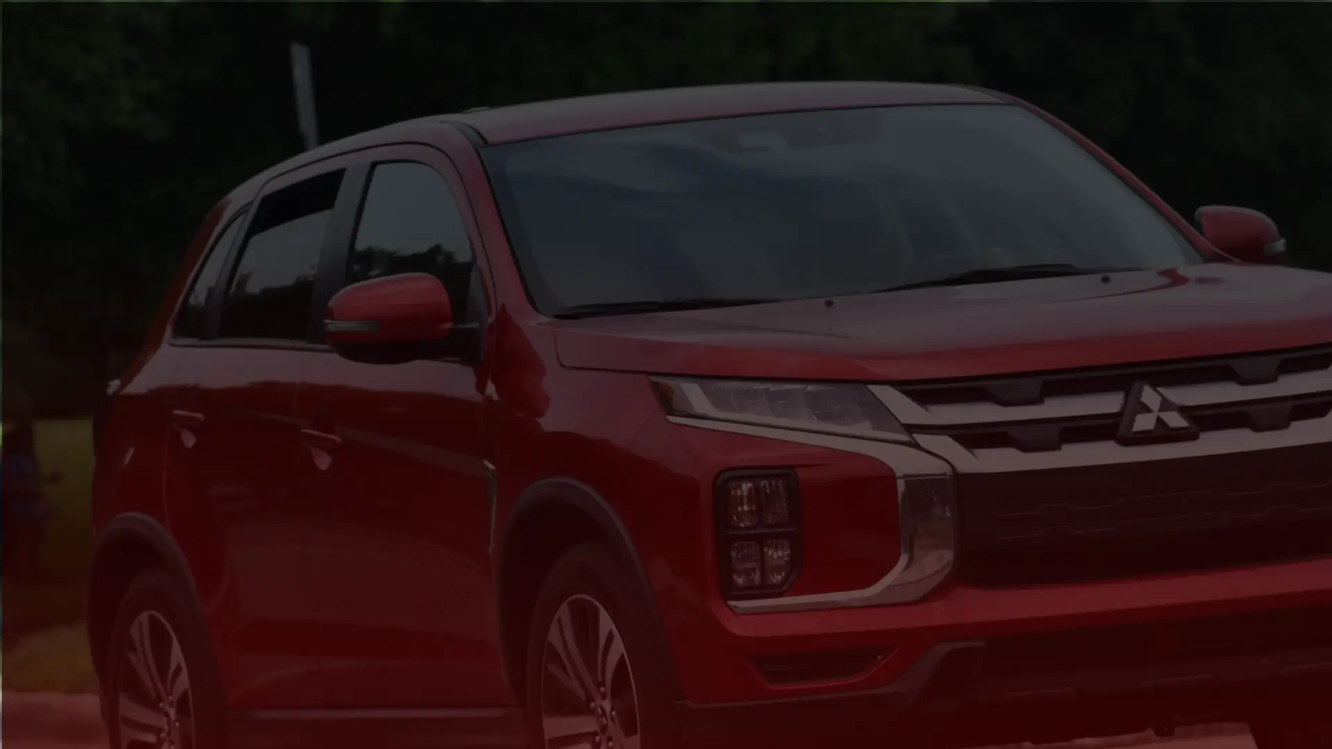 A red Mitsubishi SUV is parked outdoors, awaiting its next adventure around Greater Toronto. The cars design features a sleek grille, LED headlights, and side mirrors matching the body color. In the background is a blurred view of green trees under a cloudy sky. EuroMechanic | Top Reviewed Auto Repair Shop | Greater Toronto and Guelph | Visit Euromechanic