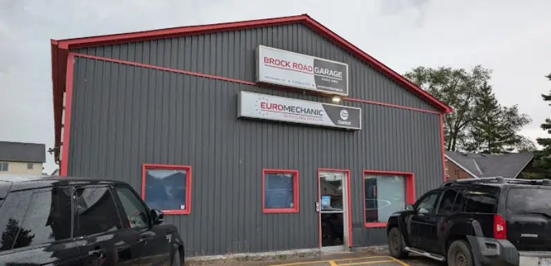 A gray metal building with red trim houses the Brock Road Garage, prominently featuring signs for Brock Road Garage and Euro Mechanic. In front, two black SUVs are parked, with trees providing a scenic backdrop to this noteworthy location. EuroMechanic | Top Reviewed Auto Repair Shop | Greater Toronto and Guelph | Visit Euromechanic
