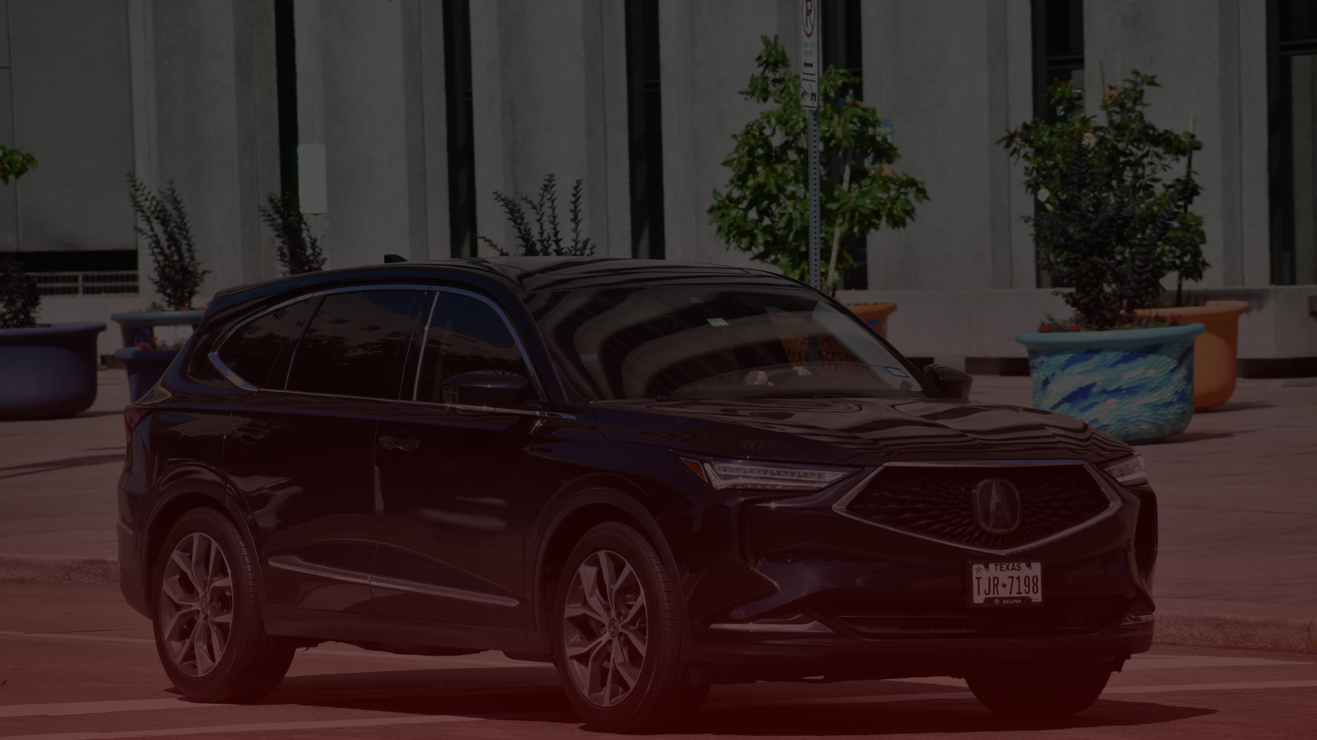 A dark-colored SUV, possibly heading to an Acura mechanic in Greater Toronto and Guelph, is parked on a city street. The vehicle sits before a modern building with large windows and planters brimming with greenery. The license plate is partially visible. EuroMechanic | Top Reviewed Auto Repair Shop | Greater Toronto and Guelph | Visit Euromechanic