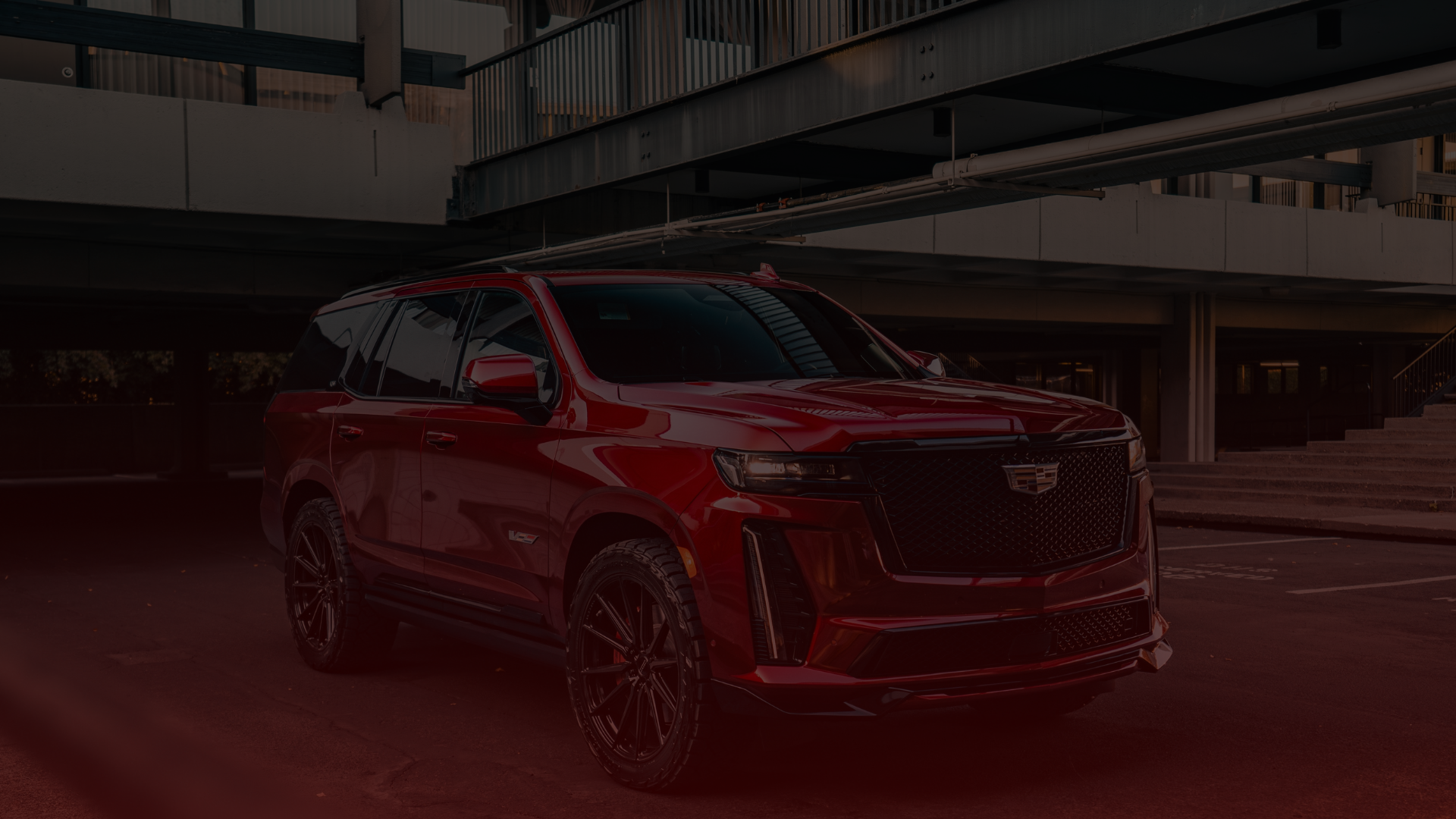 A sleek, shiny red SUV is parked in a dimly lit urban setting. Its clear a Cadillac mechanics touch can be felt in its perfection. The vehicle boasts tinted windows, black alloy wheels, and distinctive front grilles. The backdrop features industrial-style architecture reminiscent of Greater Toronto. EuroMechanic | Top Reviewed Auto Repair Shop | Greater Toronto and Guelph | Visit Euromechanic