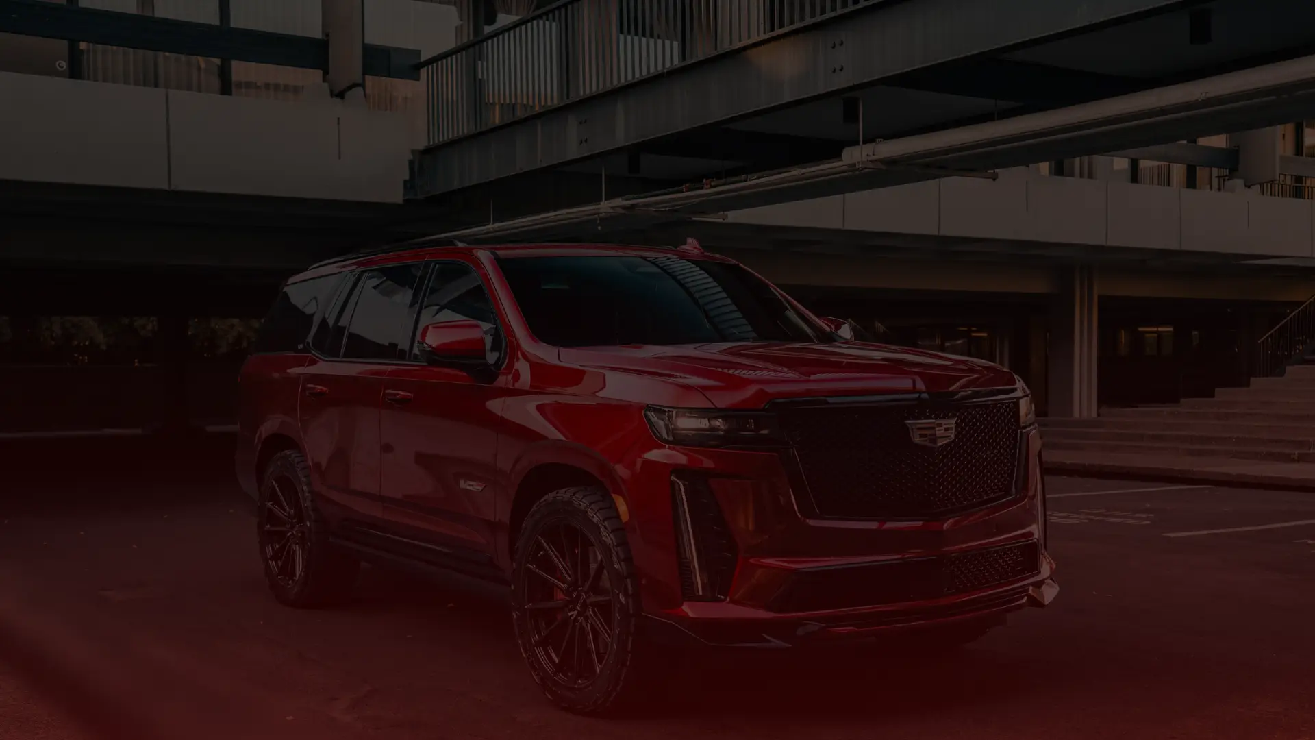 A sleek, shiny red SUV is parked in a dimly lit urban setting. Its clear a Cadillac mechanics touch can be felt in its perfection. The vehicle boasts tinted windows, black alloy wheels, and distinctive front grilles. The backdrop features industrial-style architecture reminiscent of Greater Toronto. EuroMechanic | Top Reviewed Auto Repair Shop | Greater Toronto and Guelph | Visit Euromechanic