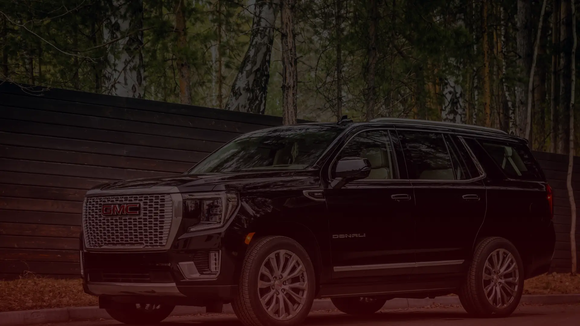 A black GMC Denali SUV is parked on a road in front of a dark wooden fence, surrounded by tall trees. The vehicle, fit for any GM mechanic in Greater Toronto, is facing front-left, proudly showcasing its grille and headlights. EuroMechanic | Top Reviewed Auto Repair Shop | Greater Toronto and Guelph | Visit Euromechanic