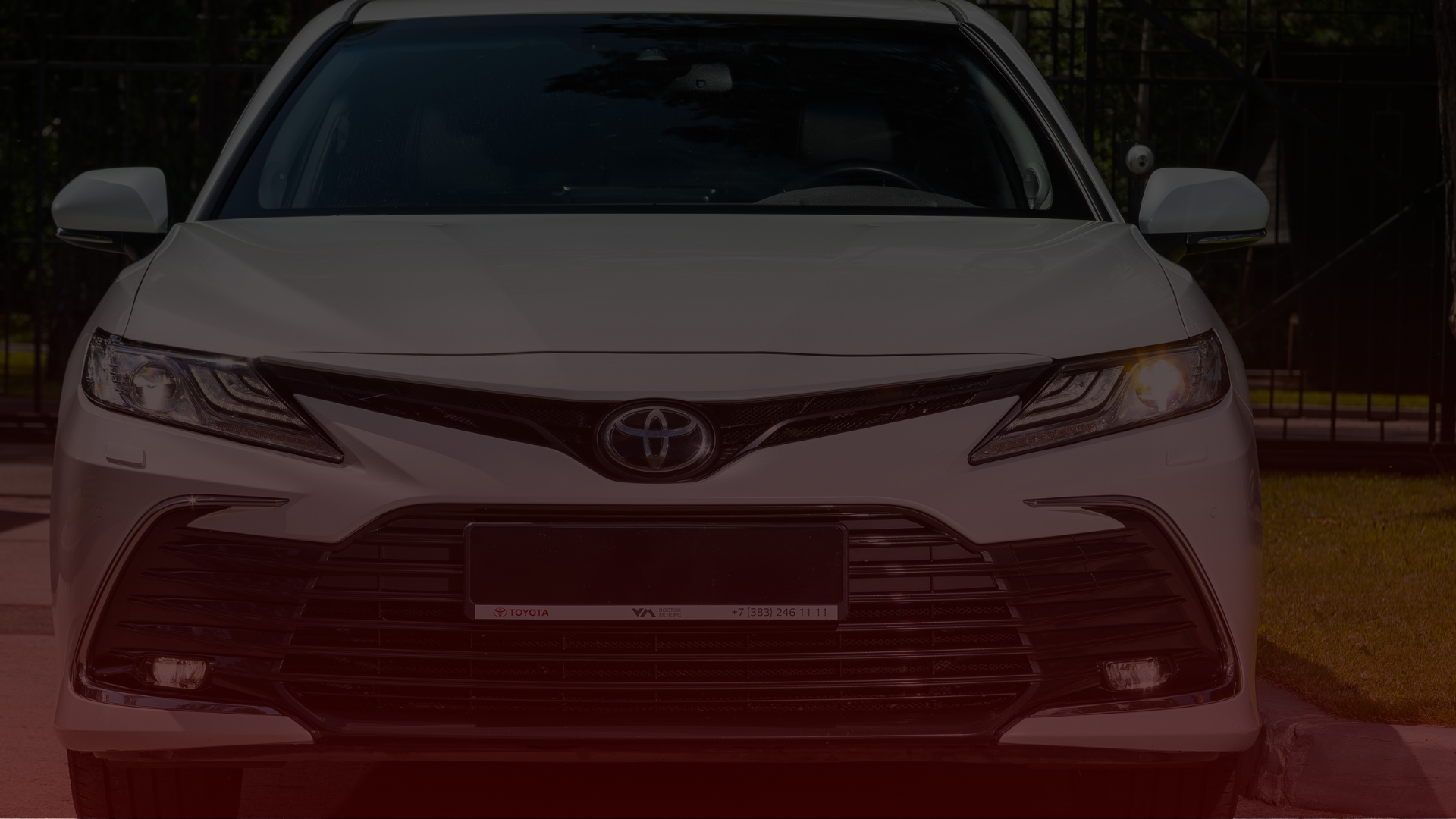 Front view of a white Toyota sedan parked outside in Greater Toronto, its headlights aglow with the emblem on the grille standing out. A backdrop of fencing and greenery complements this reliable vehicle, serviced by trusted Toyota mechanics. EuroMechanic | Top Reviewed Auto Repair Shop | Greater Toronto and Guelph | Visit Euromechanic