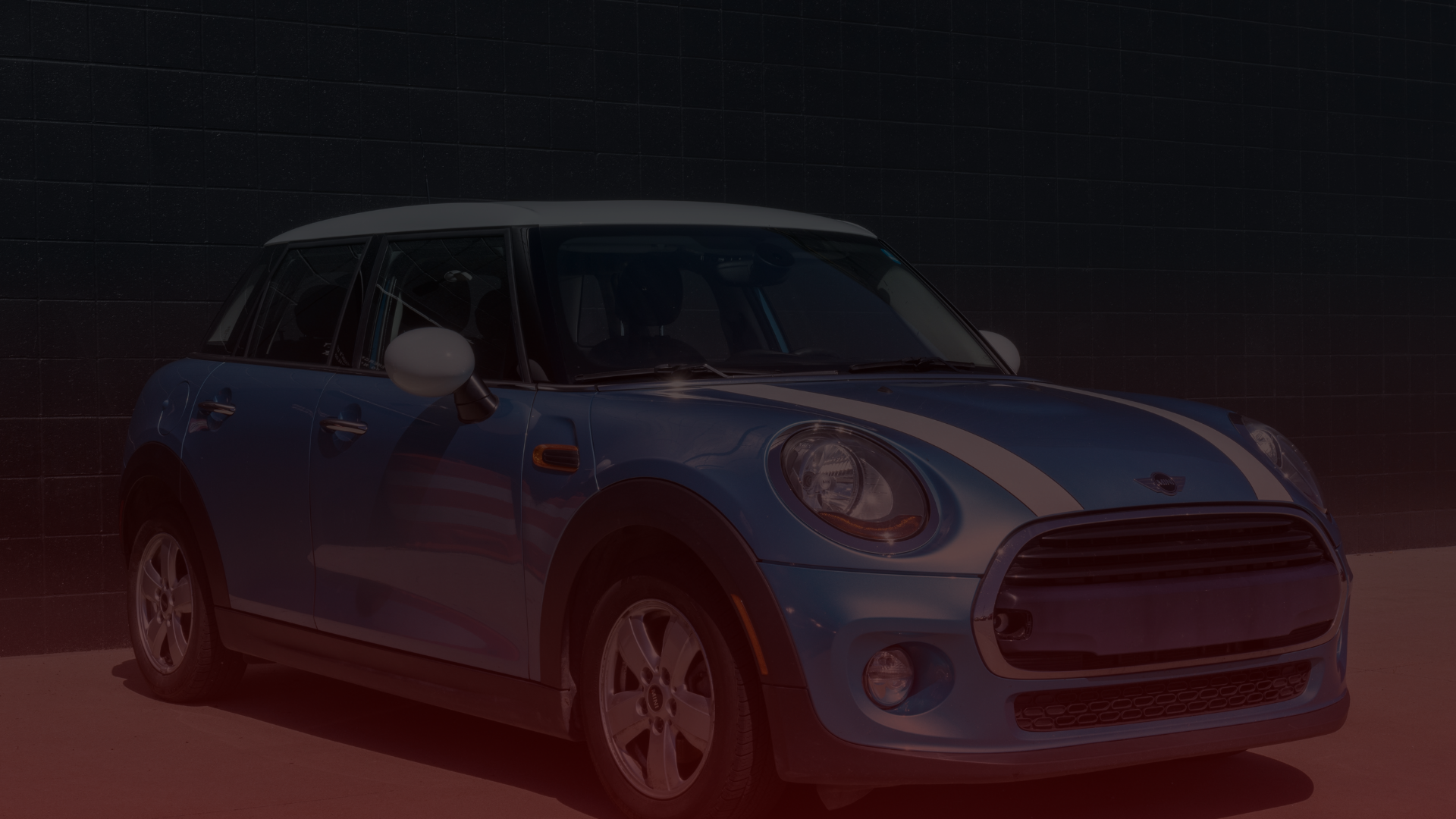A Blue Mini Cooper with white racing stripes stands in front of a dark brick wall in the Greater Toronto and Guelph area. The car sports four doors, silver rims, a white roof, and side mirrors. Its perfectly illuminated for a clear view of its front and side. EuroMechanic | Top Reviewed Auto Repair Shop | Greater Toronto and Guelph | Visit Euromechanic
