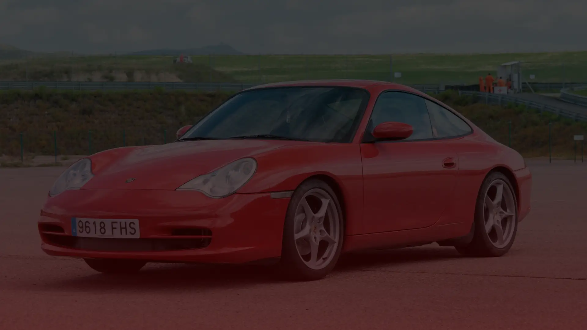 A red sports car, possibly a Porsche, is parked on an open road before a green hilly landscape under an overcast sky. In the distance near a fence, a few people gather, perhaps discussing their route back to Greater Toronto. EuroMechanic | Top Reviewed Auto Repair Shop | Greater Toronto and Guelph | Visit Euromechanic